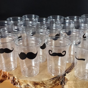 May include: A group of clear plastic cups with black mustache designs. The cups are arranged in a semi-circle on a wooden surface.