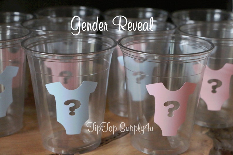 May include: Clear plastic cups with blue and pink gender reveal stickers. The stickers feature a baby onesie with a question mark. The text "Gender Reveal" is written above the cups.