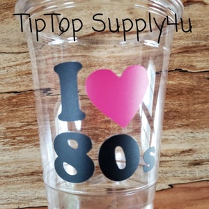 May include: Clear plastic cup with the text "I &hearts; 80s" in black and a pink heart. The words "TipTop Supply4u" are printed in black at the top of the cup. The cup is on a wooden surface.