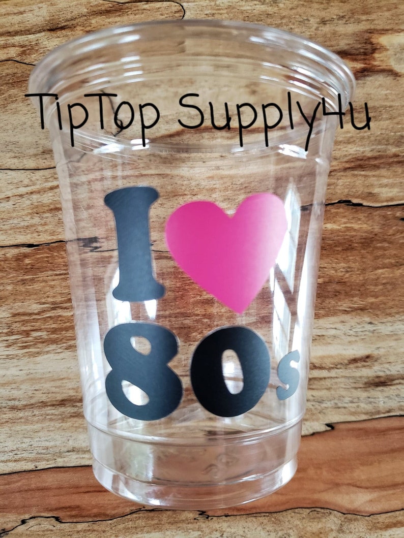 May include: Clear plastic cup with the text "I &hearts; 80s" in black and a pink heart. The words "TipTop Supply4u" are printed in black at the top of the cup. The cup is on a wooden surface.