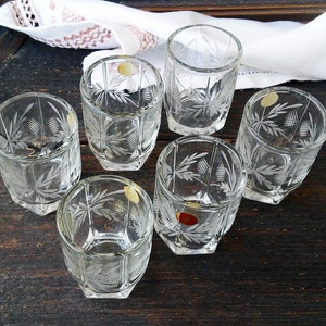 Puede incluir: Seis vasos de chupito de vidrio transparente con un diseño de vidrio tallado. Los vasos están dispuestos en círculo sobre una superficie de madera oscura.