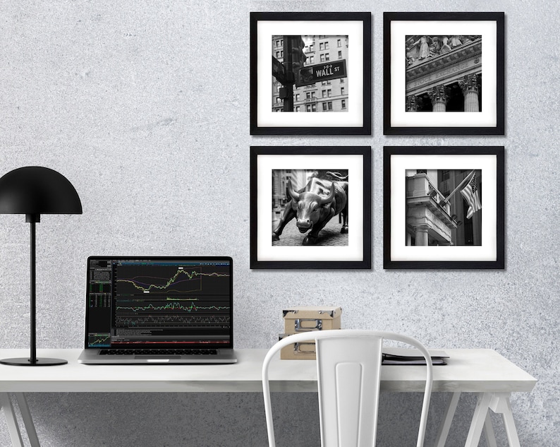 May include: A white desk with a black lamp, a laptop, and a box on the left side. Four black framed photos of New York City landmarks hang on a grey wall behind the desk. The photos show Wall Street, the Charging Bull statue, and the New York Stock Exchange.