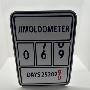 May include: A rectangular sign with the text "JIMOLDOMETER" at the top. Below are three boxes displaying the numbers 0, 6, and 9. The bottom of the sign reads "DAYS 2520290". The sign has a black border and white background.