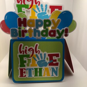 May include: A pop-up birthday card with the words "Happy Birthday!" and "high FIVE ETHAN" in colorful letters. The card features balloons and handprints, with a green, blue, and red color scheme.
