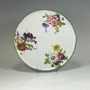May include: A white ceramic plate with a green rim features a floral design. The plate is decorated with colorful flowers, including pink, purple, yellow, and white. The flowers are arranged in a symmetrical pattern around the center of the plate.