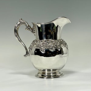 May include: A silver pitcher with an ornate floral design and the inscription "Florence" on the side. The pitcher has a rounded body and a flared spout.