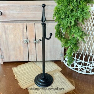 May include: A black metal coat rack with a decorative top and a curved hook. The rack stands on a round base and is displayed on a wooden surface with vintage sheet music. A green plant and a white wicker basket are in the background.