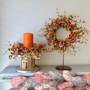 May include: A bright orange pillar candle sits in a wooden pedestal holder surrounded by a wreath of faux autumn leaves and red berries. Beside it, a matching wreath is displayed on a metal stand. A garland of muted pink and grey leaves hangs below.