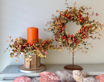 May include: A bright orange pillar candle sits in a wooden pedestal holder surrounded by a wreath of faux autumn leaves and red berries. Beside it, a matching wreath is displayed on a metal stand. A garland of muted pink and grey leaves hangs below.