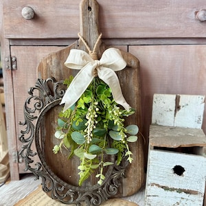 May include: A rustic wooden cutting board with a metal frame and a sprig of greenery tied with a white ribbon. The board is hanging on a light pink cabinet.