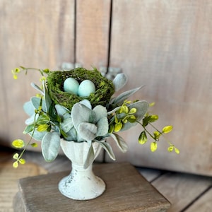 May include: A small white ceramic urn with a nest of moss and two blue eggs on top. The urn is sitting on a wooden pedestal with a carved base.