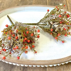 May include: Two sprigs of faux greenery with small red berries. The sprigs are on a white oval tray with a brown wooden rim.