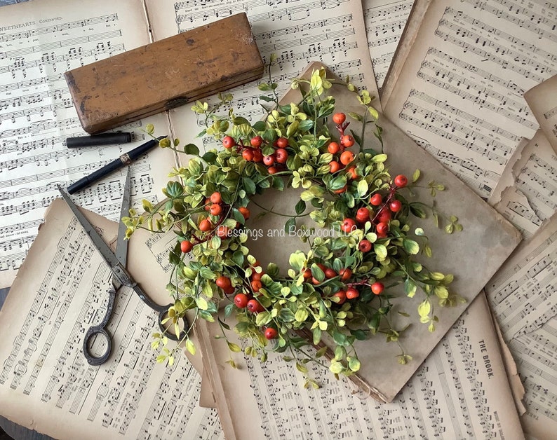 May include: A circular wreath featuring green leaves and small orange berries, positioned on vintage sheet music. A wooden box, a black pen, and a pair of metal scissors are placed nearby. The text Blessings and Boxwood LLC is visible.