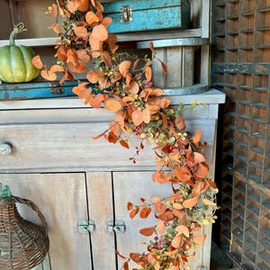 Peut inclure: Une guirlande d&eacute;corative compos&eacute;e de branches d'eucalyptus artificielles aux feuilles rondes dans des tons orange br&ucirc;l&eacute;, rouille et vert doux. La structure flexible en forme de vigne est drap&eacute;e verticalement, pr&eacute;sentant un arrangement dense de feuillage aux teintes et textures vari&eacute;es inspir&eacute;es de l'automne sur toute la longueur de la pi&egrave;ce.