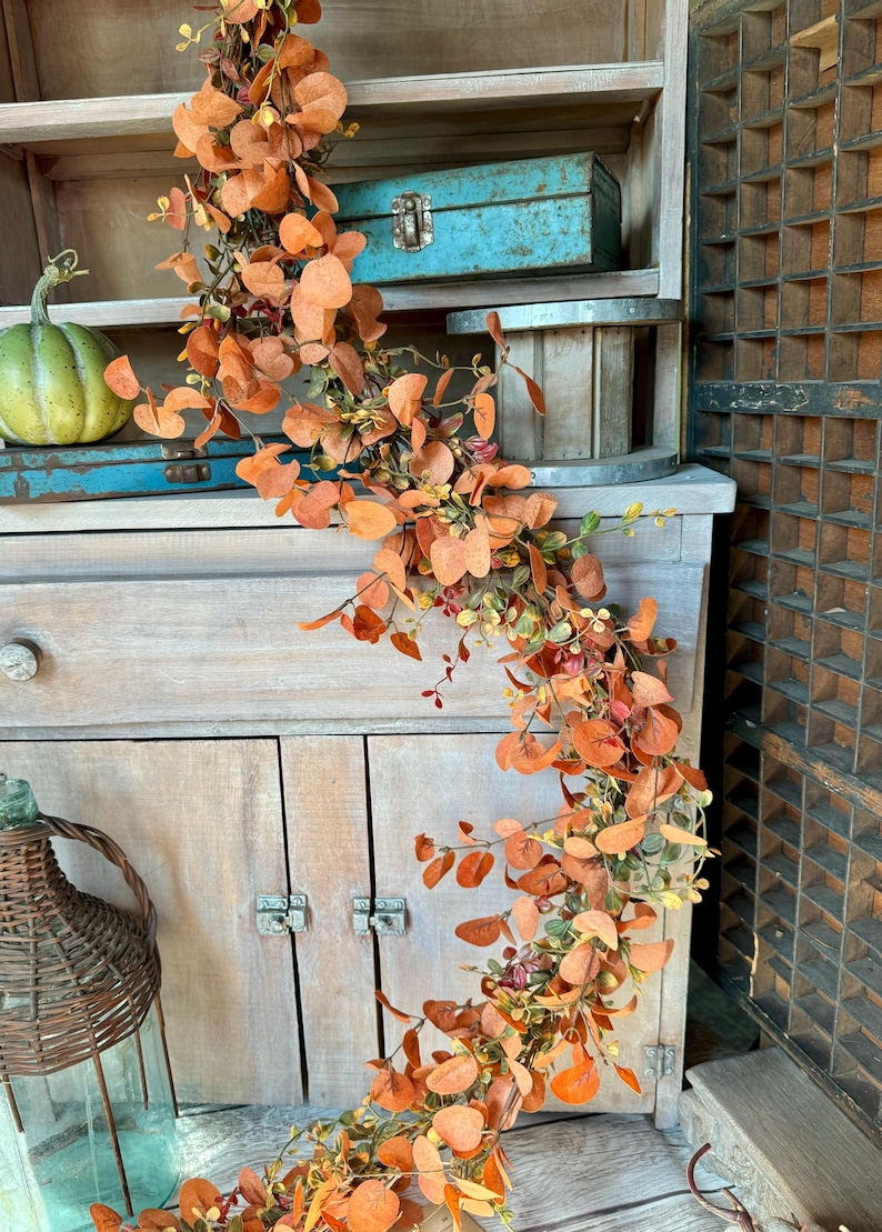 Peut inclure: Une guirlande d&eacute;corative compos&eacute;e de branches d'eucalyptus artificielles aux feuilles rondes dans des tons orange br&ucirc;l&eacute;, rouille et vert doux. La structure flexible en forme de vigne est drap&eacute;e verticalement, pr&eacute;sentant un arrangement dense de feuillage aux teintes et textures vari&eacute;es inspir&eacute;es de l'automne sur toute la longueur de la pi&egrave;ce.