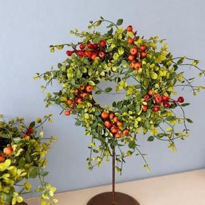 May include: A circular wreath featuring vibrant green leaves and clusters of small, bright red berries. The wreath is mounted on a thin, dark brown metal stand with a circular base, displaying a natural aesthetic with thin, branching stems.