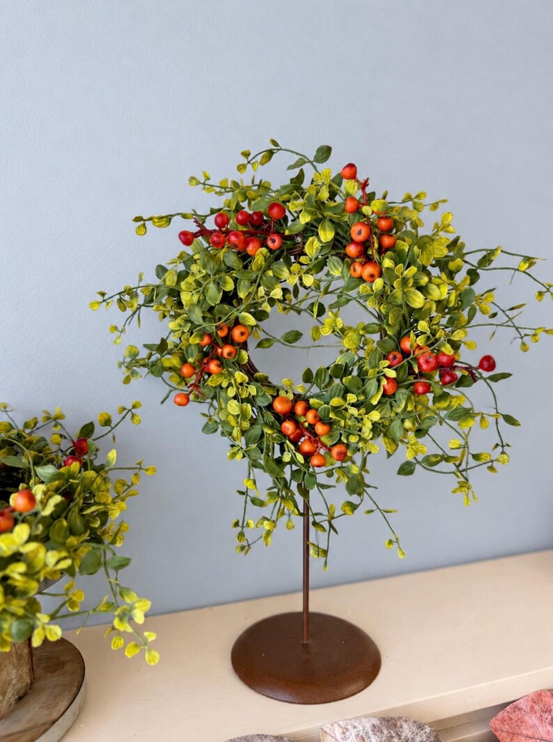 May include: A circular wreath featuring vibrant green leaves and clusters of small, bright red berries. The wreath is mounted on a thin, dark brown metal stand with a circular base, displaying a natural aesthetic with thin, branching stems.