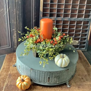 May include: A cylindrical orange pillar candle sits inside a wooden dough bowl filled with green faux boxwood foliage and orange berries. A small white pumpkin and a small orange pumpkin are placed on a round, gray, fluted metal pedestal.