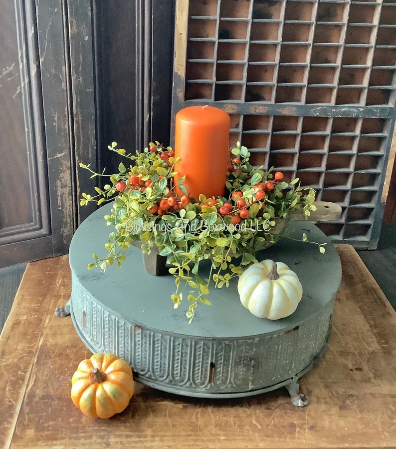 May include: A cylindrical orange pillar candle sits inside a wooden dough bowl filled with green faux boxwood foliage and orange berries. A small white pumpkin and a small orange pumpkin are placed on a round, gray, fluted metal pedestal.
