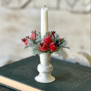 May include: A white candle sits in a decorative, distressed white candle holder. The holder is adorned with red berries, green pine needles, and silver leaves. The candle holder is placed on a dark green book, creating a festive holiday display.
