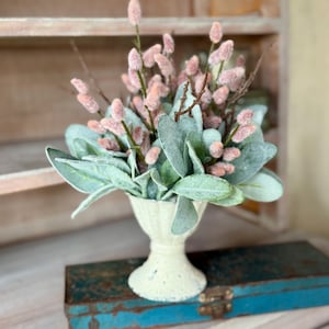 May include: A small white ceramic vase with a decorative design holds a bouquet of faux dusty miller and pink fluffy flowers.