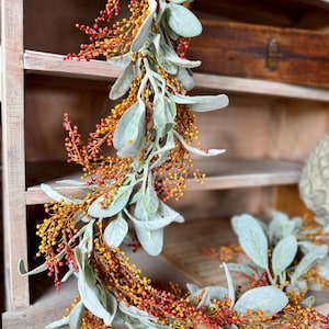 May include: A decorative garland featuring sage green leaves and clusters of small, orange and red berries. The garland is arranged in a circular shape, with the leaves and berries intertwined. The background is a wooden shelf.