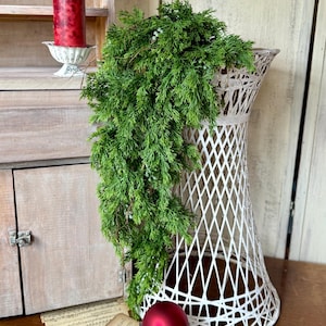 May include: A tall, white wicker plant stand holds a cascade of green foliage. A red Christmas ornament sits on a decorative stand. A red candle sits on a decorative candle holder. Vintage sheet music is scattered around.
