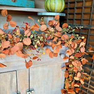 May include: A decorative garland featuring autumn-colored eucalyptus leaves in shades of orange, rust, and muted green, draped over a light-colored wooden cabinet. A small, speckled green pumpkin sits on a metal stand above the foliage.