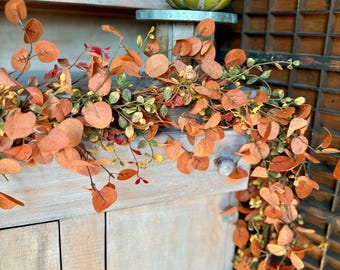 May include: A decorative garland featuring autumn-colored eucalyptus leaves in shades of orange, rust, and muted green, draped over a light-colored wooden cabinet. A small, speckled green pumpkin sits on a metal stand above the foliage.