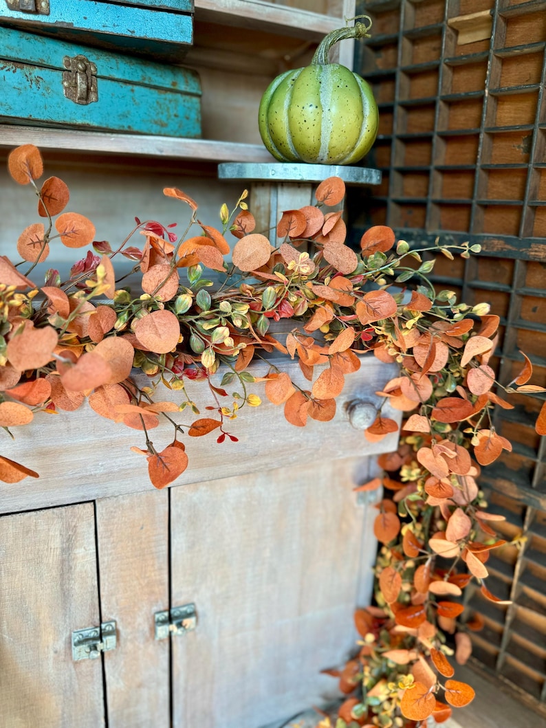 Peut inclure: Une guirlande d&eacute;corative compos&eacute;e de feuilles d'eucalyptus aux couleurs automnales, dans des tons orange, rouille et vert doux, drap&eacute;e sur un meuble en bois clair. Une petite citrouille verte tachet&eacute;e repose sur un support en m&eacute;tal.