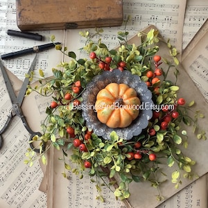 May include: A small orange pumpkin sits in the center of a fluted gray metal tart pan, surrounded by a wreath of green leaves and small orange berries. The arrangement is placed atop vintage sheet music with a wooden box, pen, and scissors.