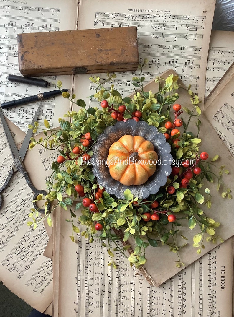 May include: A small orange pumpkin sits in the center of a fluted gray metal tart pan, surrounded by a wreath of green leaves and small orange berries. The arrangement is placed atop vintage sheet music with a wooden box, pen, and scissors.