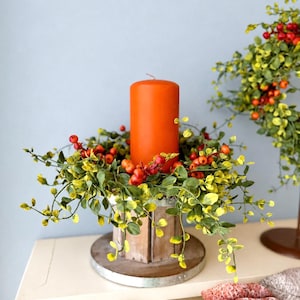 May include: A bright orange pillar candle sits inside a circular wooden candle holder base. The candle is surrounded by a decorative ring of artificial green leaves and small red berries. A matching wreath with green leaves and red berries is visible.