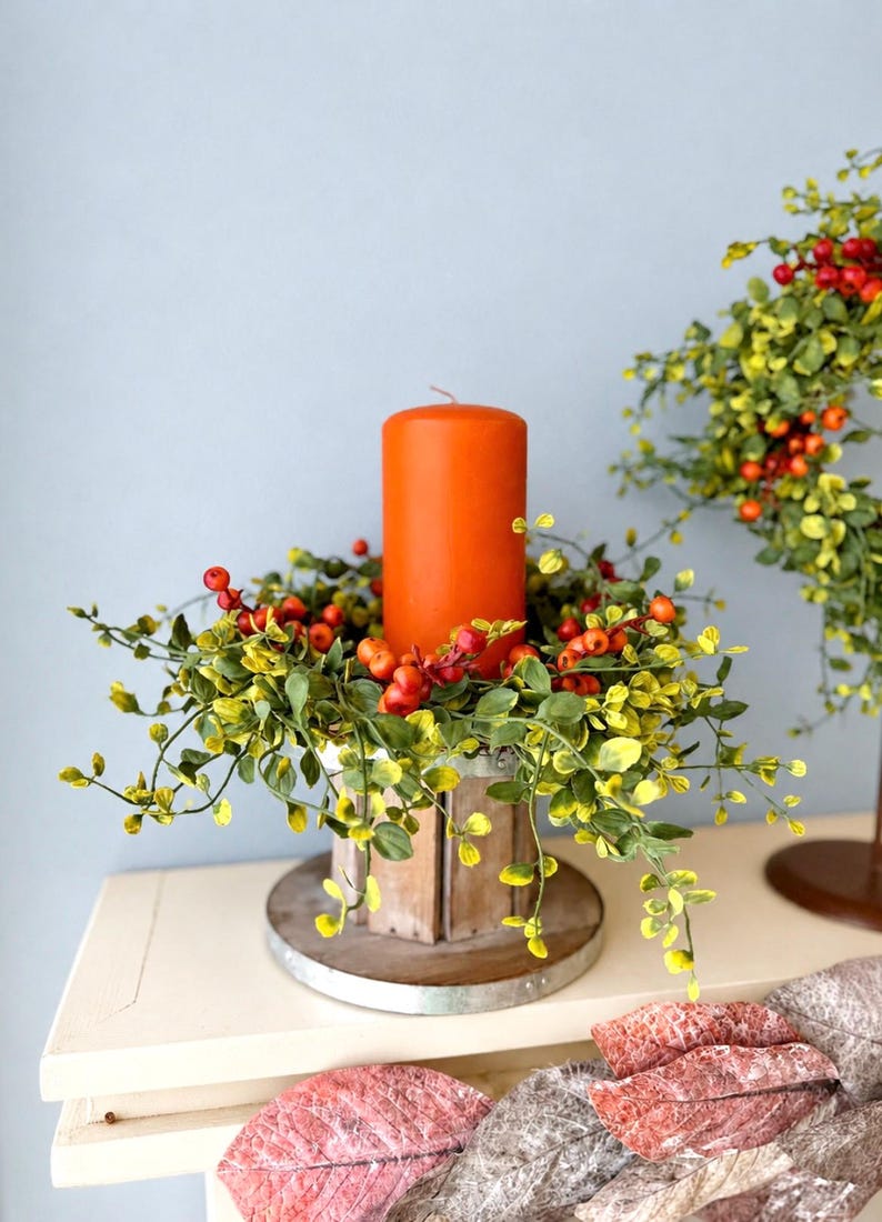 May include: A bright orange pillar candle sits inside a circular wooden candle holder base. The candle is surrounded by a decorative ring of artificial green leaves and small red berries. A matching wreath with green leaves and red berries is visible.