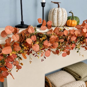 Peut inclure: Une guirlande artificielle compos&eacute;e de feuilles d'eucalyptus rondes couleur rouille et de petits accents jaunes. La guirlande est drap&eacute;e sur une chemin&eacute;e blanche, accompagn&eacute;e de deux bougeoirs noirs et de trois citrouilles d&eacute;coratives blanches, vertes et orange.