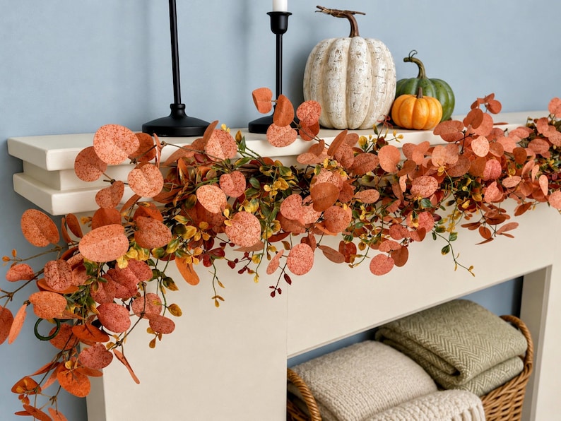 Peut inclure: Une guirlande artificielle compos&eacute;e de feuilles d'eucalyptus rondes couleur rouille et de petits accents jaunes. La guirlande est drap&eacute;e sur une chemin&eacute;e blanche, accompagn&eacute;e de deux bougeoirs noirs et de trois citrouilles d&eacute;coratives blanches, vertes et orange.