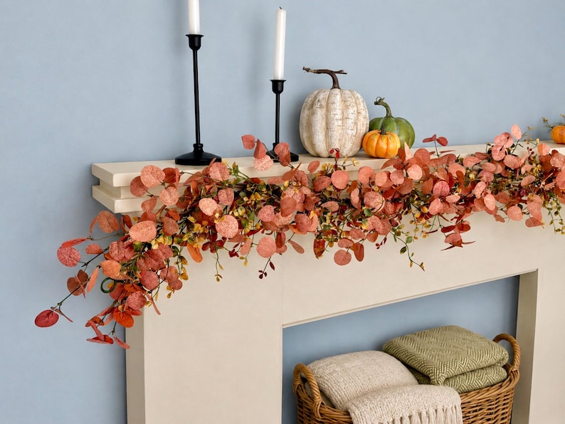 May include: A decorative garland featuring round, rust-colored eucalyptus leaves and small green sprigs draped across a mantel. Two black metal candle holders with white candles, a white pumpkin, and a green pumpkin sit on the mantel.