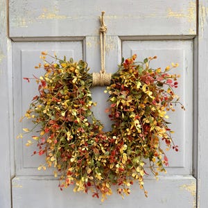 May include: A circular wreath made of colorful faux foliage, featuring green, yellow, and red leaves. The wreath is suspended by a natural rope loop, tied with a knot. The wreath is hanging on a weathered, light blue door.