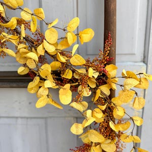 May include: A decorative garland featuring yellow, round leaves and small red berries. The garland is arranged on a brown stem, creating a natural and autumnal aesthetic. The background is a light gray surface.
