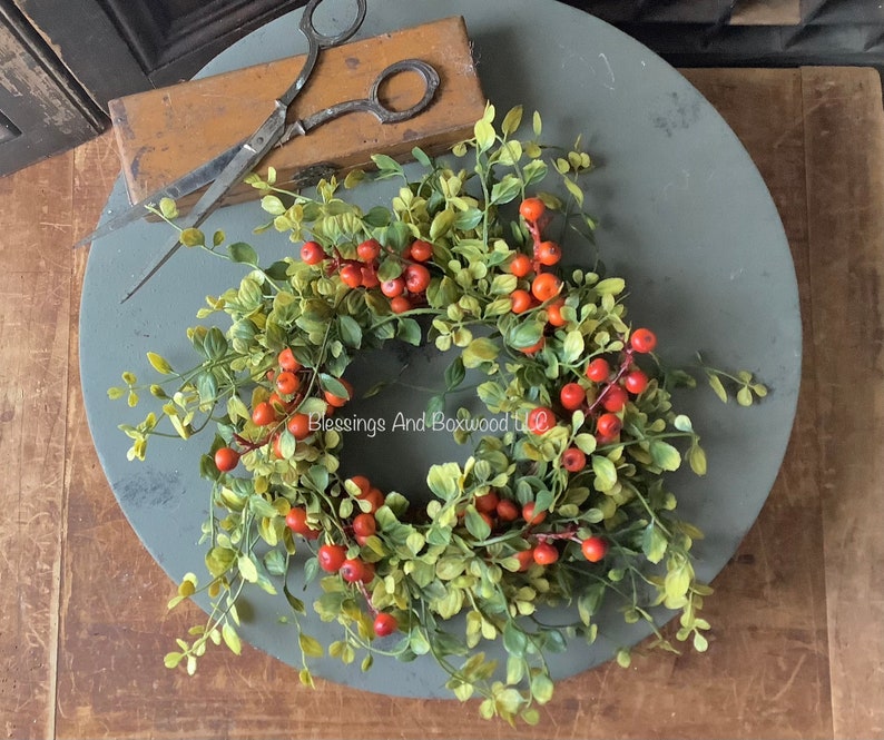 May include: A green and red artificial wreath with red berries and green leaves. The wreath is on a gray round surface. The text 'Blessings And Boxwood LLC' is visible in the center of the wreath.