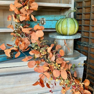 Peut inclure: Une guirlande d&eacute;corative compos&eacute;e de nombreuses feuilles rondes aux tons orang&eacute;s sur de fines tiges brunes, drap&eacute;e &agrave; c&ocirc;t&eacute; d'une citrouille ronde vert clair avec des taches sombres pos&eacute;e sur un petit socle m&eacute;tallique circulaire.