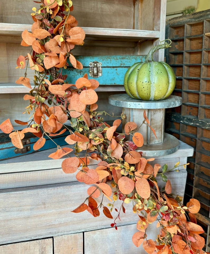 Peut inclure: Une guirlande d&eacute;corative compos&eacute;e de nombreuses feuilles rondes aux tons orang&eacute;s sur de fines tiges brunes, drap&eacute;e &agrave; c&ocirc;t&eacute; d'une citrouille ronde vert clair avec des taches sombres pos&eacute;e sur un petit socle m&eacute;tallique circulaire.