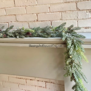 Peut inclure: Une guirlande de pin givr&eacute;e avec des baies rouges drap&eacute; sur une chemin&eacute;e blanche. La guirlande est d&eacute;cor&eacute;e de pommes de pin et a un aspect enneig&eacute; et hivernal. Le texte "BlessingsAndBoxwood.Etsy.com" est visible sur la chemin&eacute;e.