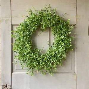 May include: A round, green wreath made of small, leafy sprigs. The wreath is hanging on a weathered, light gray door. The wreath is a vibrant green color, and the door has a distressed, vintage look.