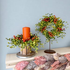 May include: A bright orange pillar candle sits inside a wooden crate ringed with green leaves and red berries. Beside it, a matching wreath of green leaves and red berries stands on a metal pedestal. A garland of large, textured, multi-colored leaves.