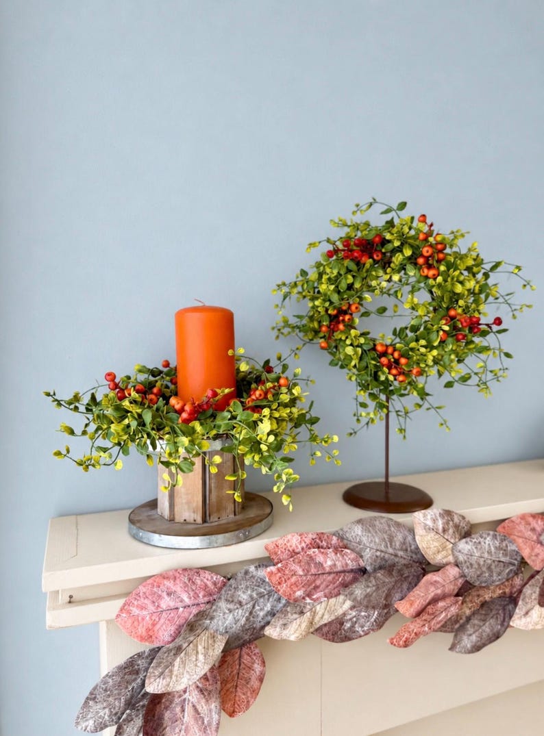May include: A bright orange pillar candle sits inside a wooden crate ringed with green leaves and red berries. Beside it, a matching wreath of green leaves and red berries stands on a metal pedestal. A garland of large, textured, multi-colored leaves.