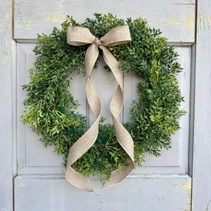 May include: A round, green wreath made of leafy foliage, adorned with a beige burlap bow and flowing ribbon. The wreath is a decorative item, perfect for adding a touch of nature to any home decor.