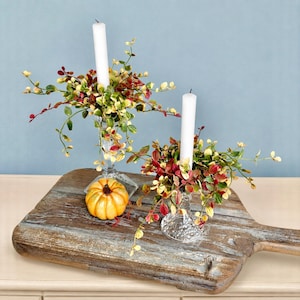 May include: Two clear glass candle holders each containing a white taper candle and surrounded by a ring of artificial foliage with green, yellow, and red leaves. A small, orange, ribbed decorative pumpkin sits beside the candle holders.