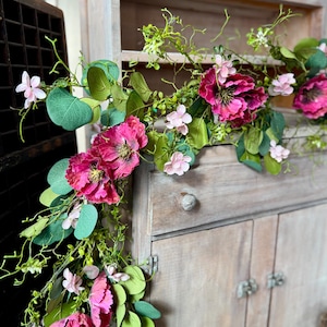 May include: A floral garland with large, vibrant pink peonies, small pink flowers, and green eucalyptus leaves. The garland is draped over a rustic, light-colored wooden cabinet with a distressed finish, creating a charming, vintage aesthetic.