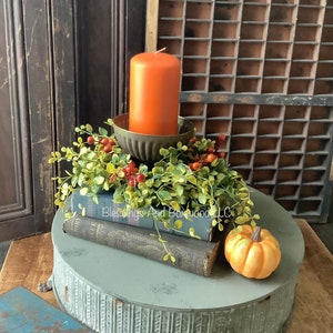 May include: A burnt orange pillar candle sits in a fluted metal holder atop two stacked vintage books. A faux greenery wreath with small red berries encircles the candle base. A small orange pumpkin rests nearby on a round, gray metal pedestal stand.